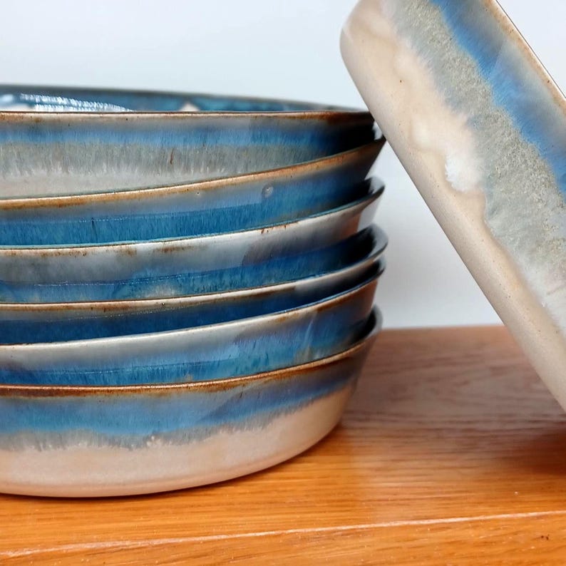 May include: A stack of ceramic bowls with a blue and white glaze. The bowls have a wide rim and a shallow depth. The glaze transitions from a light blue to a darker blue, with a white band near the rim. The bowls are stacked on a wooden surface.