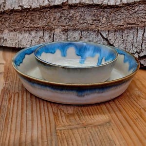 May include: A ceramic bowl set with a white base and a blue glaze that drips down the sides. The set includes a large, shallow bowl and a smaller bowl nested inside. The bowls are on a wooden surface.
