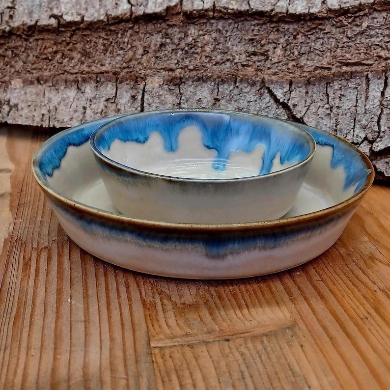 May include: A ceramic bowl set with a white base and a blue glaze that drips down the sides. The set includes a large, shallow bowl and a smaller bowl nested inside. The bowls are on a wooden surface.