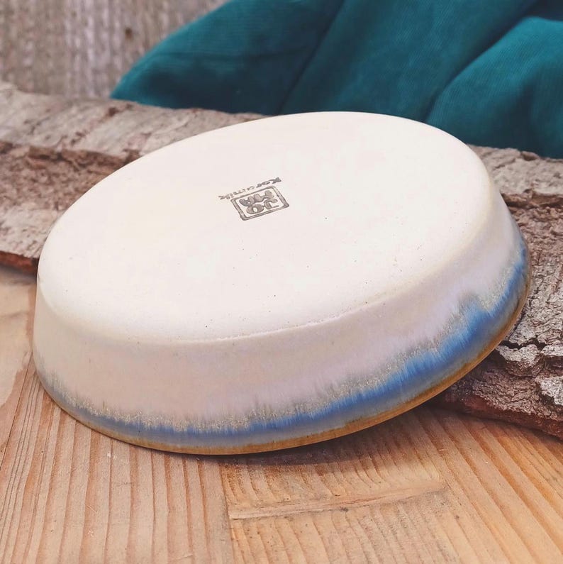 May include: A handmade ceramic bowl with a white interior and a blue-glazed rim. The bowl has a slightly oval shape and sits on a wooden surface. The bottom of the bowl has a stamped logo.