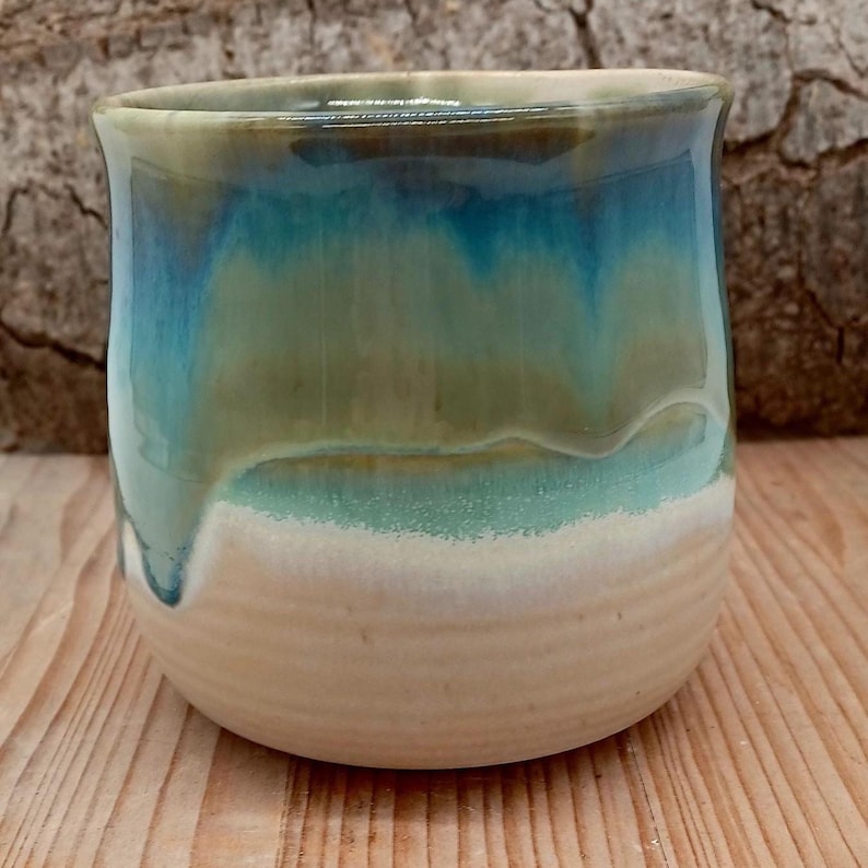 Puede incluir: Un vaso de cer&aacute;mica hecho a mano con un esmalte &uacute;nico. La parte superior del vaso presenta una mezcla de tonos azules y verdes que gotean sobre una base blanca. La taza est&aacute; sobre una superficie de madera.