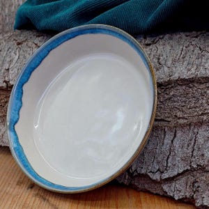 May include: A handmade ceramic serving dish with a white interior and a vibrant blue rim. The dish has a slightly irregular shape and a natural, earthy aesthetic. The rim is painted with a bright blue glaze, adding a pop of color to the neutral tones.