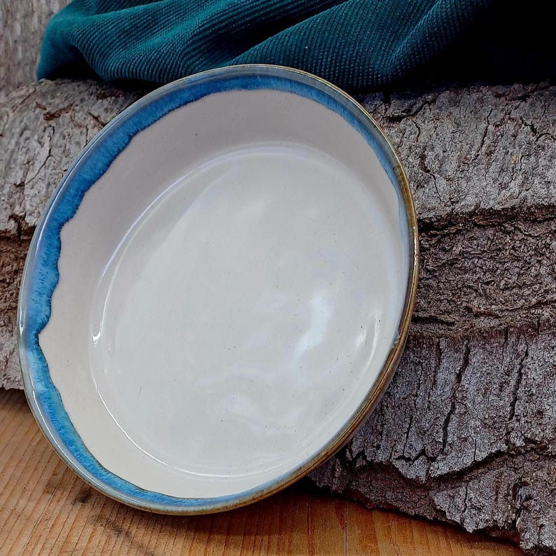 May include: A handmade ceramic serving dish with a white interior and a vibrant blue rim. The dish has a slightly irregular shape and a natural, earthy aesthetic. The rim is painted with a bright blue glaze, adding a pop of color to the neutral tones.