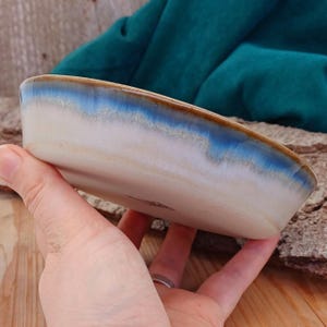 May include: A handmade ceramic dish with a rectangular shape. The dish features a cream-colored base with a blue and brown rim. The glaze has a glossy finish, and the dish is held in a person's hand.
