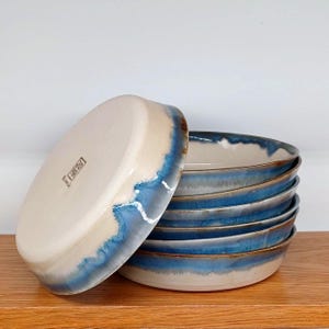 May include: A stack of handmade ceramic bowls with a unique design. The bowls have a cream-colored interior and a blue and white glazed rim. The bowls are stacked on a wooden surface. The bowls are likely used for serving food.