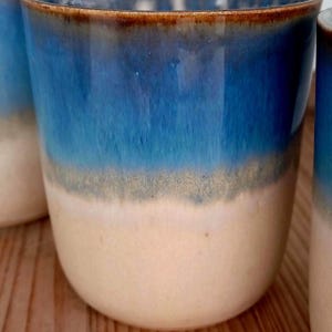Op de afbeelding: Een set keramische mokken met een gradi&euml;ntontwerp. De mokken lopen over van lichtbeige aan de basis naar diepblauw aan de bovenkant, met daartussen een grijsgroene band. De rand is donkerbruin.