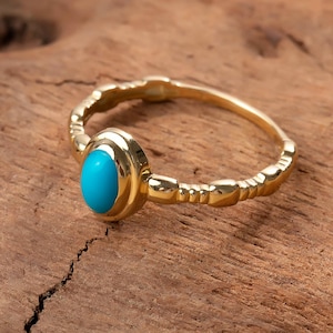 May include: A gold ring featuring a turquoise gemstone. The ring has a textured band and a bezel setting for the oval, bright blue stone. The ring is displayed on a piece of brown wood.
