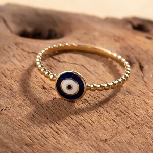 May include: A gold-colored ring with a beaded band and a circular charm. The charm features a blue background with a white and black eye design. The ring is set against a wooden surface.
