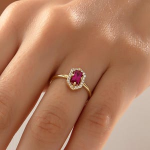 May include: A gold ring with a rectangular, deep red gemstone encircled by small, clear stones. The ring sits on a slender gold band, displaying a classic and elegant design. The ring is shown on a hand.