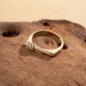 May include: A gold-colored ring with a unique geometric design. The ring features a square shape with a rounded inner band. A small section of the ring is adorned with multiple small, clear gemstones. The ring is displayed on a wooden surface.