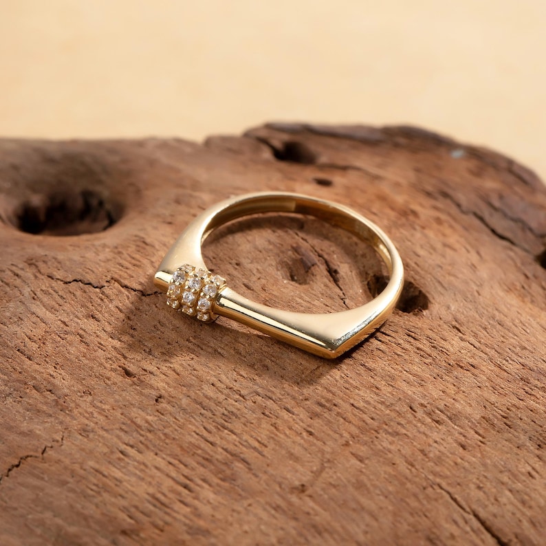May include: A gold-colored ring with a unique geometric design. The ring features a square shape with a rounded inner band. A small section of the ring is adorned with multiple small, clear gemstones. The ring is displayed on a wooden surface.