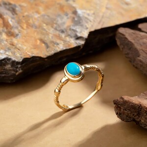 May include: A gold ring featuring a turquoise oval gemstone. The ring's band has a textured design. The turquoise stone is set in a gold bezel. The ring is displayed against a neutral backdrop.