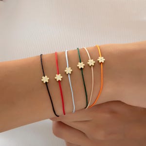 May include: A collection of daisy charm bracelets in various colors, including black, red, white, green, beige, and orange. Each bracelet features a delicate gold-colored daisy charm. The bracelets are displayed on a person's wrist.