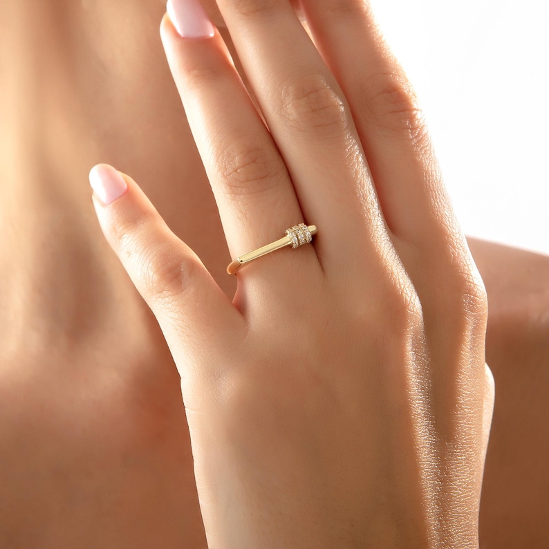 May include: A gold ring with a rectangular design and small, clear stones. The ring is worn on a finger, showcasing its delicate and elegant style. The ring's design is simple yet eye-catching, perfect for everyday wear.