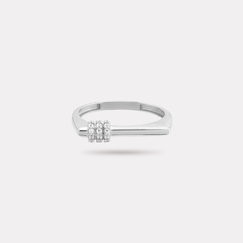 May include: A silver ring with a rectangular band and a horizontal bar design. Three small, round, clear stones are set in a vertical row on the bar. The ring has a minimalist and modern aesthetic.