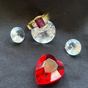May include: A gold ring with a large red gemstone and smaller clear gemstones. The ring is displayed on a black fabric surface with several clear gemstones.