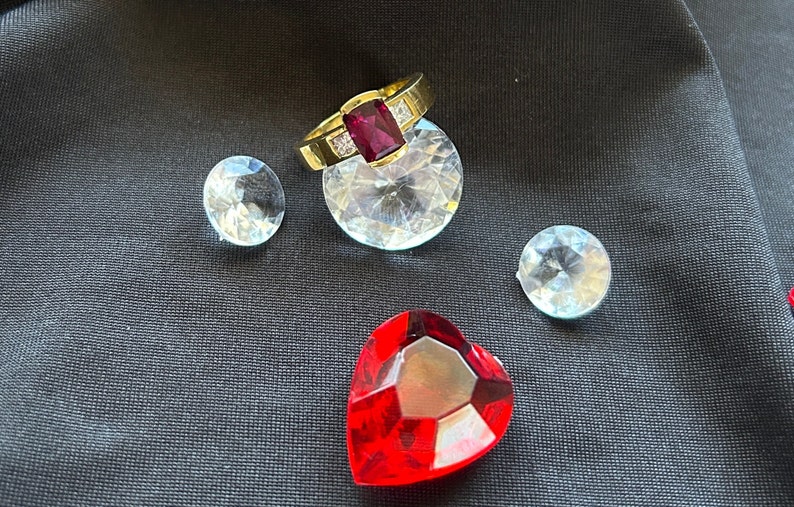 May include: A gold ring with a large red gemstone and smaller clear gemstones. The ring is displayed on a black fabric surface with several clear gemstones.