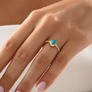 May include: A gold ring with a bright turquoise oval gemstone. The ring has a textured band with a delicate, braided pattern. The ring is displayed on a finger.