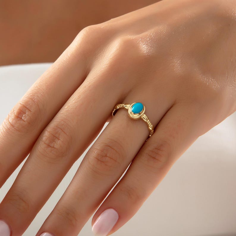 May include: A gold ring with a bright turquoise oval gemstone. The ring has a textured band with a delicate, braided pattern. The ring is displayed on a finger.