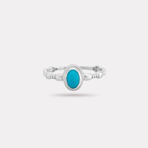 May include: A silver ring with a bright turquoise oval stone at the centre. The band has a beaded design. The ring is set against a plain white background, highlighting its elegant simplicity.