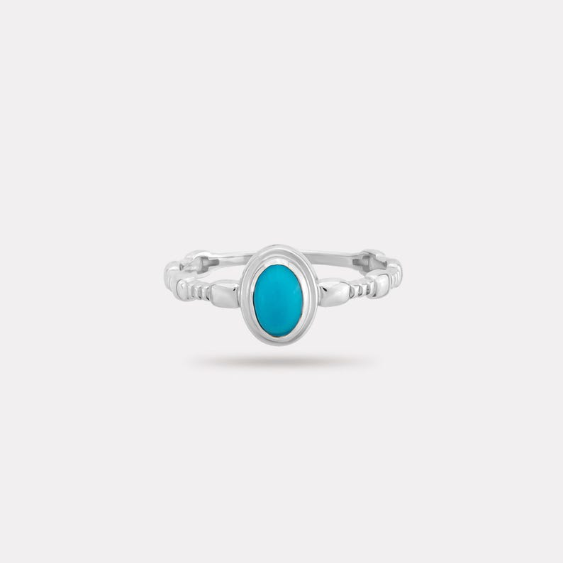 May include: A silver ring with a bright turquoise oval stone at the centre. The band has a beaded design. The ring is set against a plain white background, highlighting its elegant simplicity.
