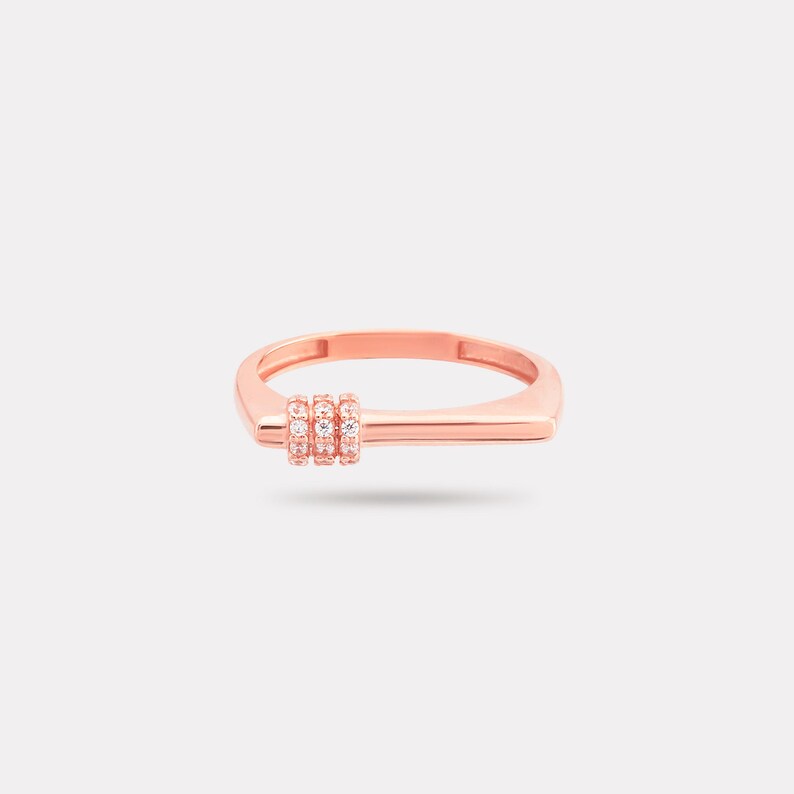 May include: Rose gold ring with a rectangular band and a cluster of three small, clear gemstones. The ring has a modern, minimalist design and is set against a plain white background.
