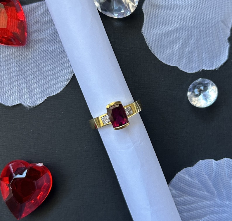 May include: A gold ring with a large, rectangular, dark red gemstone and two smaller, square, clear gemstones. The ring is on a white surface.