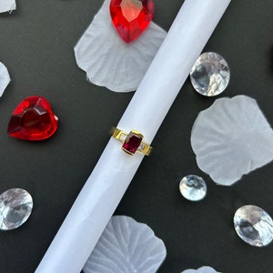 May include: A gold ring with a red gemstone set in a square shape. The ring is surrounded by white gemstones. The ring is on a white surface with red hearts and clear gemstones.