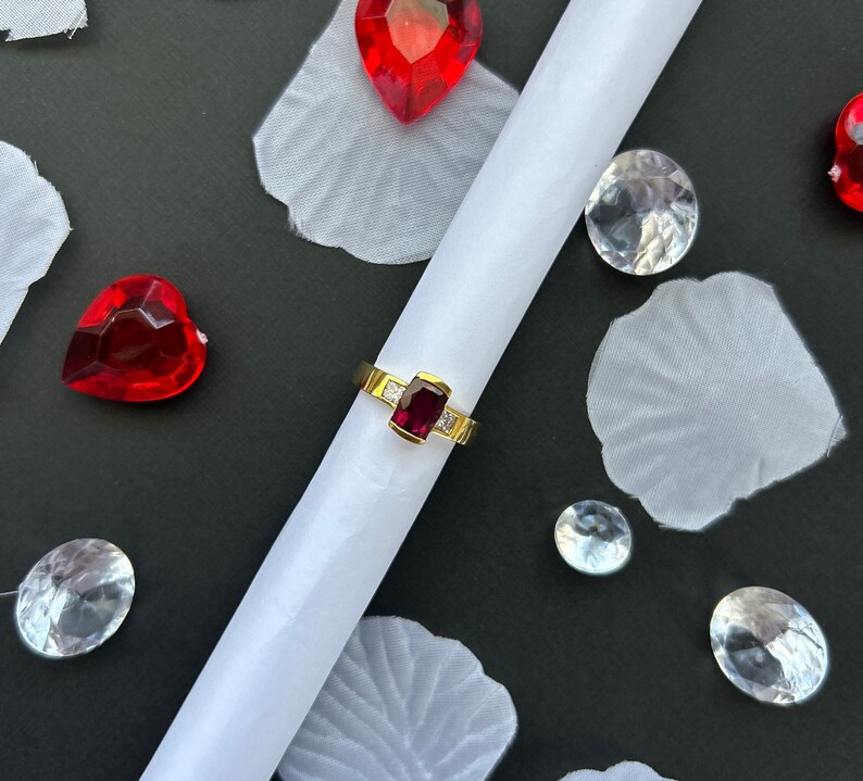 May include: A gold ring with a red gemstone set in a square shape. The ring is surrounded by white gemstones. The ring is on a white surface with red hearts and clear gemstones.