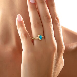 May include: A gold ring with a bright turquoise oval gemstone. The ring has a textured band and a simple bezel setting. The ring is displayed on a finger, showcasing the jewellery's design and colour.