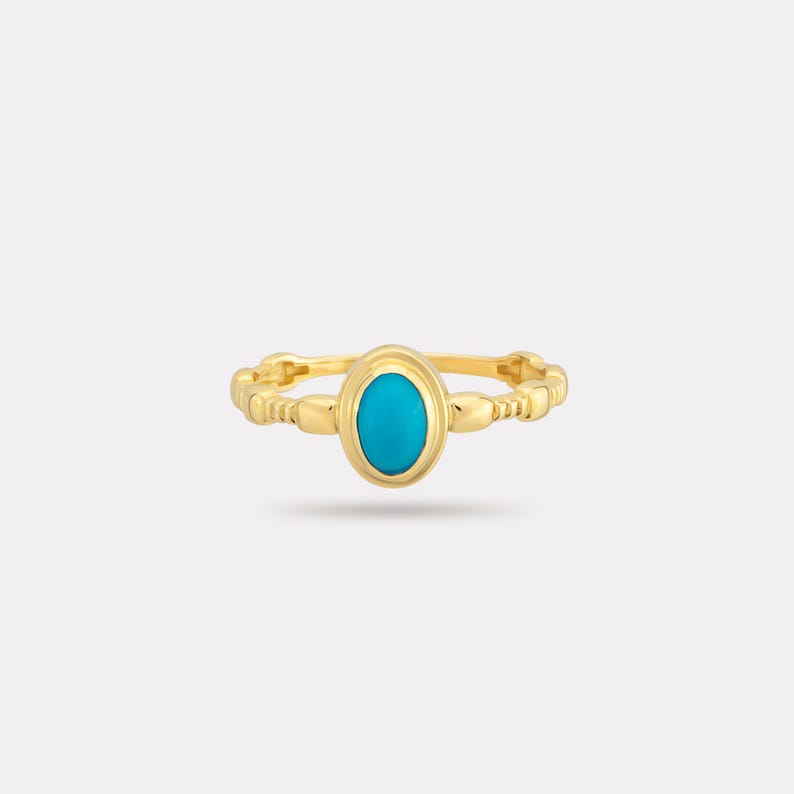 May include: A gold ring with a bright turquoise oval stone. The band has a textured design with small, repeating details. The ring is set against a white background, highlighting the jewellery's design and colour.
