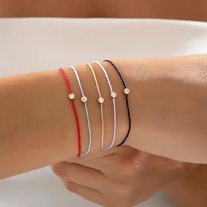 May include: A selection of fine bracelets with thin cords in red, light blue, beige, white, and black. Each bracelet has a small, round, gold-coloured charm with a clear, sparkling stone. The bracelets are shown on a wrist.