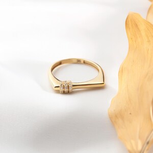 May include: A gold ring with a unique design. The ring features a rectangular band with three small, clear stones set in a row. The ring is made of shiny gold and is displayed on a white surface.