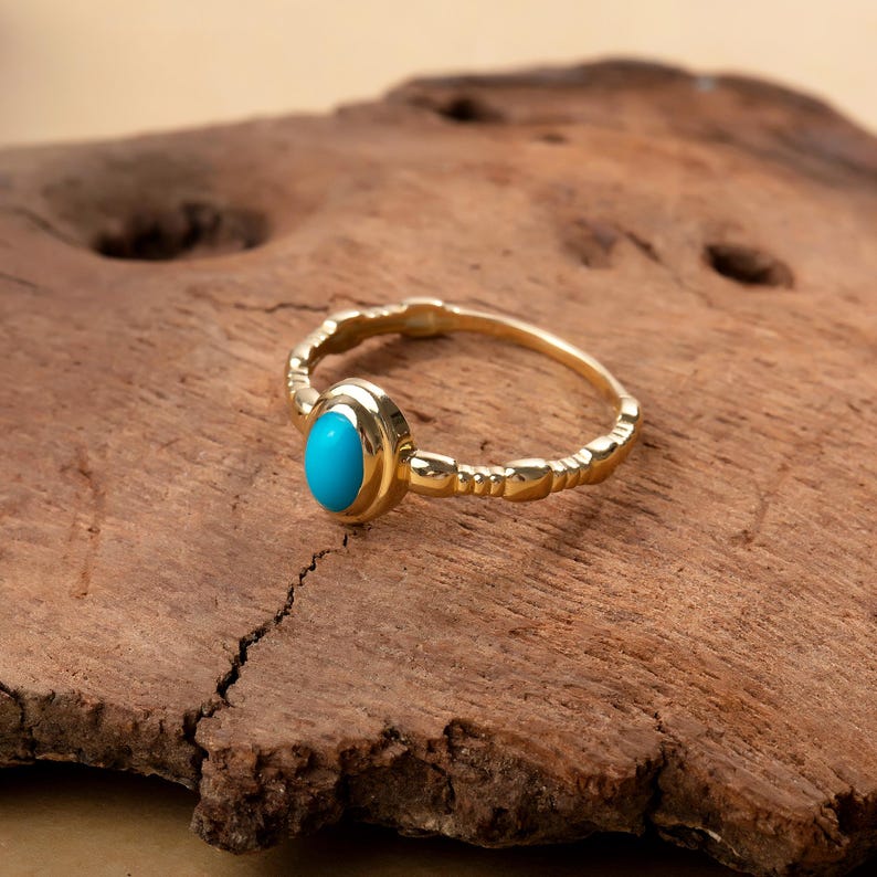 May include: A gold ring featuring a turquoise gemstone. The ring has a textured band and a bezel setting for the oval, bright blue stone. The ring is displayed on a piece of brown wood.