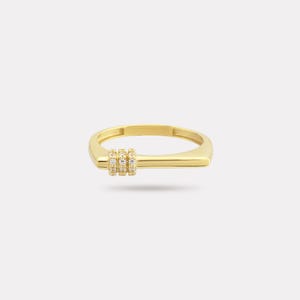 May include: A gold-colored ring with a rectangular band and a horizontal bar design. Three vertical rows of small, clear stones are set on the bar. The ring is set against a plain white background.