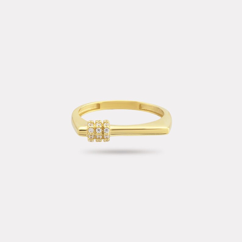 May include: A gold-colored ring with a rectangular band and a horizontal bar design. Three vertical rows of small, clear stones are set on the bar. The ring is set against a plain white background.