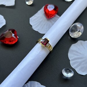 May include: A gold ring with a red gemstone and small diamonds set in the band. The ring is on a white surface with red hearts and clear crystals.