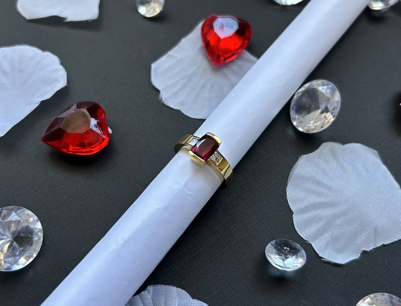 May include: A gold ring with a red gemstone and small diamonds set in the band. The ring is on a white surface with red hearts and clear crystals.