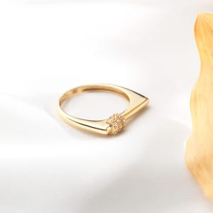 May include: A gold ring with a unique geometric design. The ring features a rectangular band that tapers to a point, with a cluster of small gemstones at the center. The ring is set against a white background.