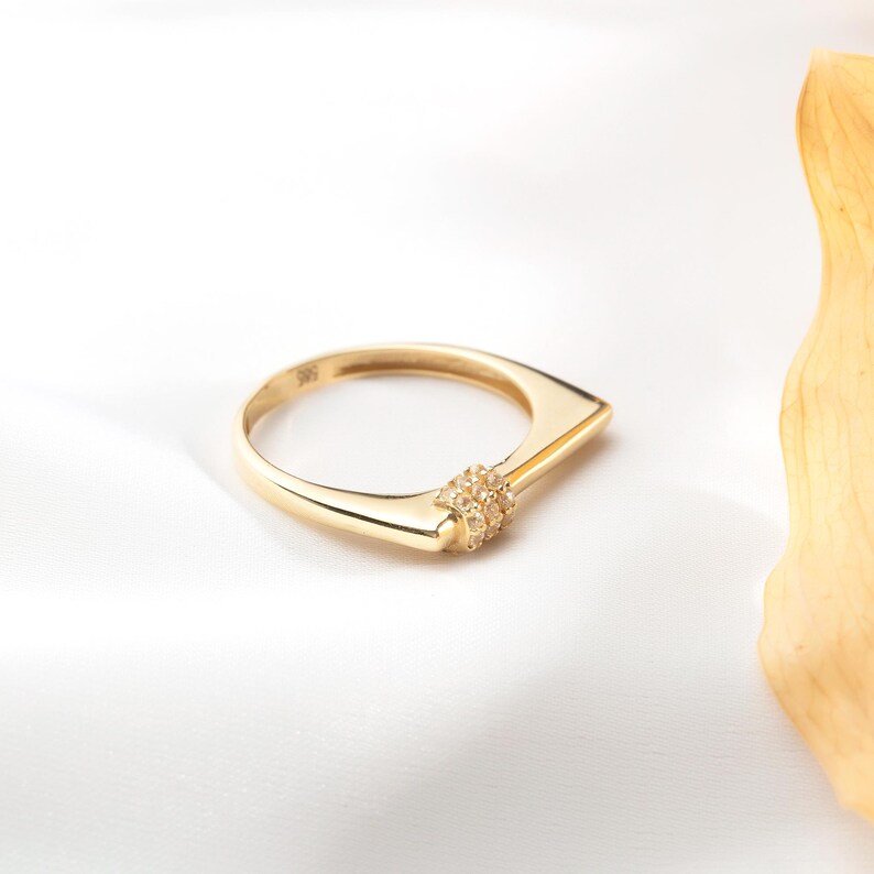 May include: A gold ring with a unique geometric design. The ring features a rectangular band that tapers to a point, with a cluster of small gemstones at the center. The ring is set against a white background.