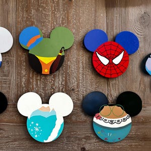 May include: A collection of colorful, wooden Mickey Mouse ear-shaped decorations. Designs include R2-D2, Spider-Man, Mickey Mouse, Elsa, Stitch, and others. The ears are painted with vibrant colors and placed against a wooden background.