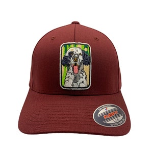 May include: A maroon baseball cap with an embroidered patch of a dog on the front. The patch is green, white, and black. The cap has a Flexfit label on the back.
