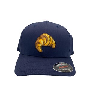 May include: A navy blue baseball cap with a yellow and brown embroidered croissant on the front. The cap has a Flexfit label on the back with the text "The Original Flexfit, 1XL, It Fits Your Life."