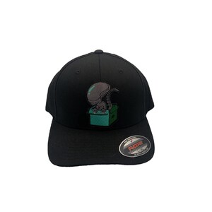 May include: Black fitted baseball cap with an embroidered design of a green alien in a green bin. The cap has a black Flexfit label on the side.