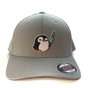 May include: Grey baseball cap featuring a cartoon penguin holding a knife. The cap has a black and red "Flexfit" logo on the back.