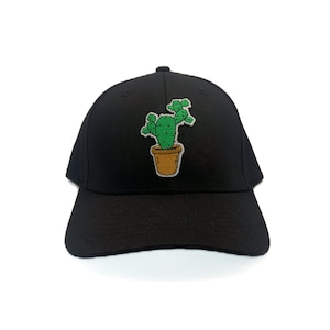 May include: Black baseball cap with an embroidered design of a green cactus in a brown pot. The cactus is detailed with green needles and sits in a light brown pot. The cap has a curved peak and a button on the crown.