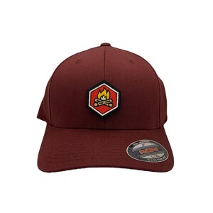 May include: A maroon baseball cap with a black hexagon patch featuring a campfire design. The patch has a red background with a yellow flame and orange logs. The cap has a black Flexfit label on the back.