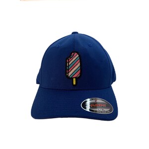 May include: A blue baseball cap with a colourful popsicle graphic embroidered on the front. The cap has a curved brim and a velcro closure. The text "Flexfit" and "1-XL" is visible on the back of the cap.