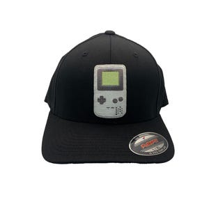 May include: Black baseball cap with a retro video game console patch. The patch features a grey handheld console with a green screen. The cap has a curved peak and a Flexfit label.