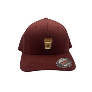 May include: A maroon baseball cap with a brown coffee cup graphic on the front. The cap has a black Flexfit label on the side.